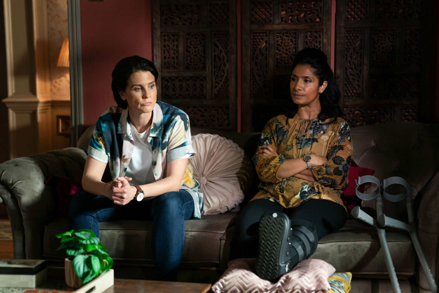 Eve and Suki sit side by side on a sofa on EastEnders