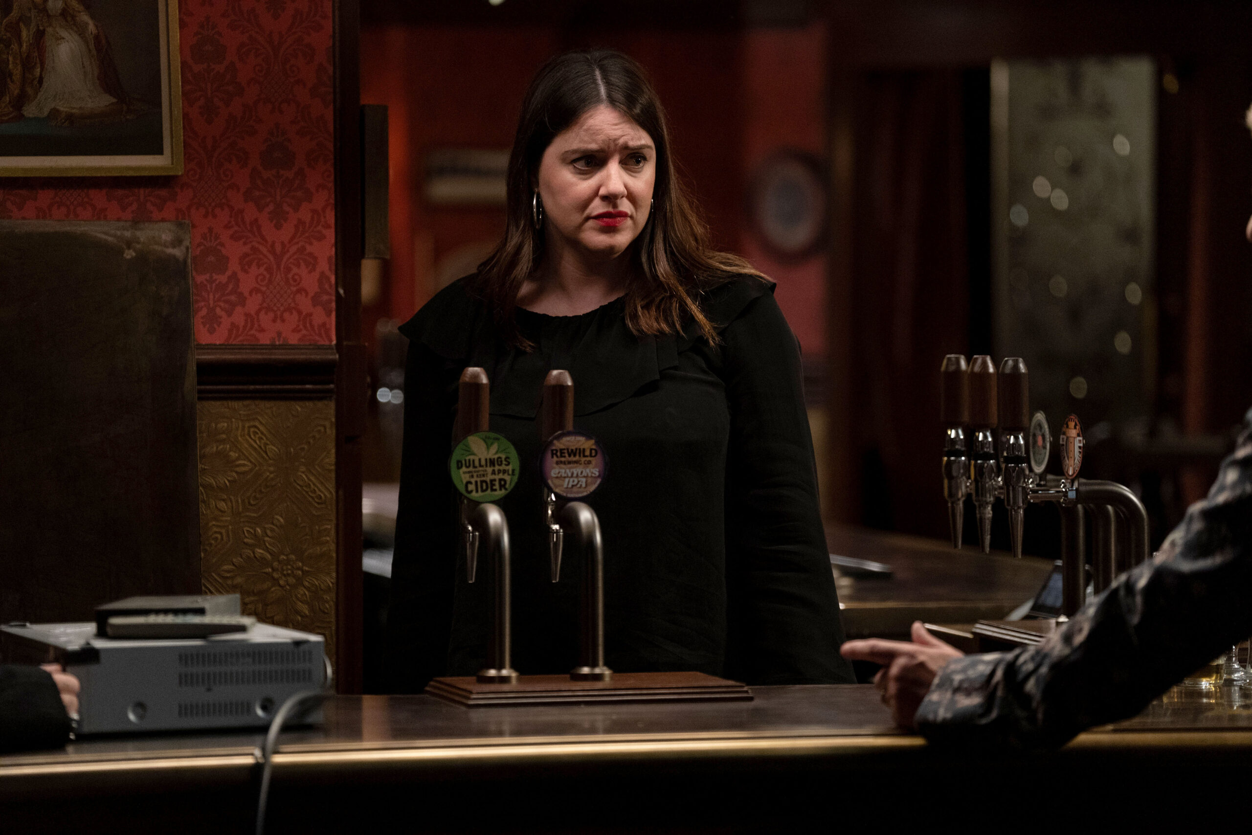 Zoe looks worried behind the bar of The Vic