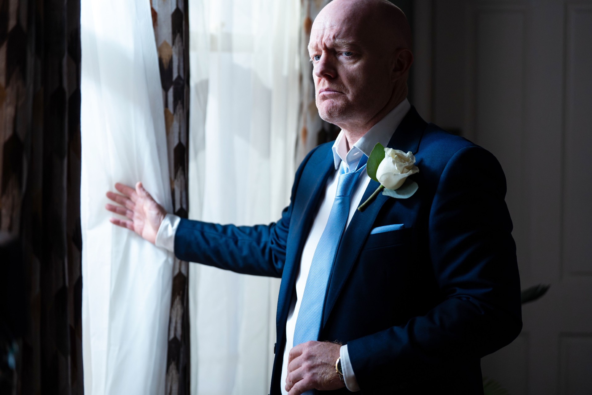 Max looks suspicious as he stands at a window in his wedding suit