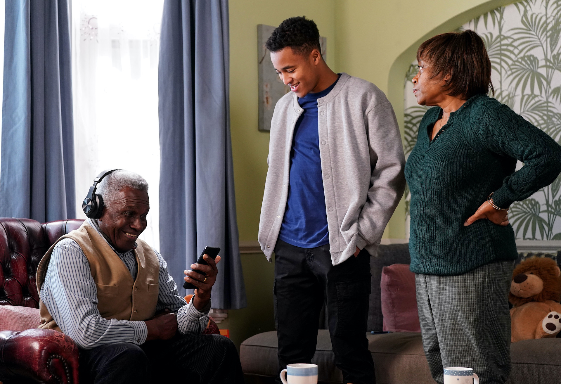 Patrick smiling while looking at a phone, as Yolande looks on concerned in EastEnders spoilers