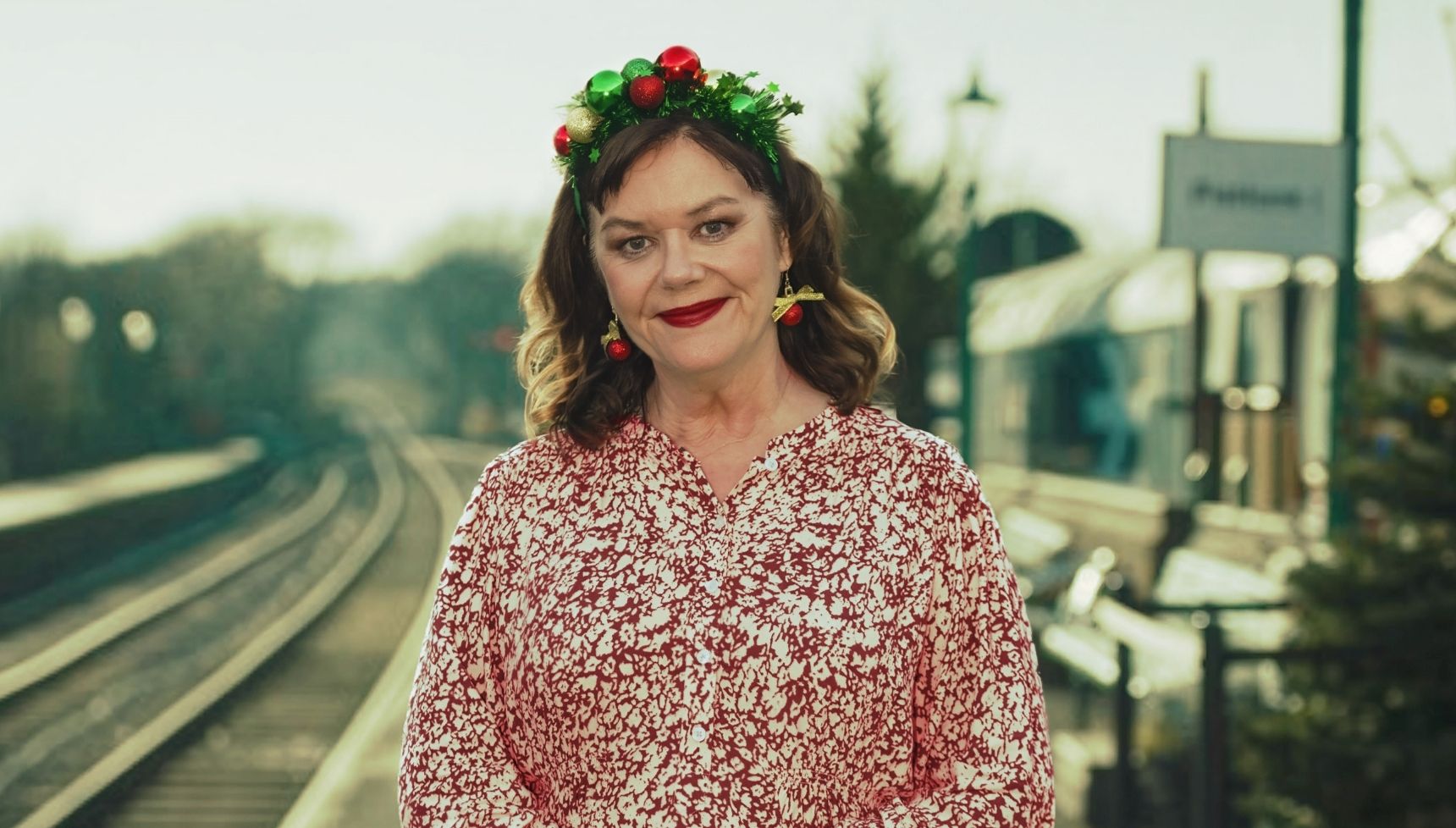 Josie Lawrence wearing a Christmas hairband and standing on a train station platform