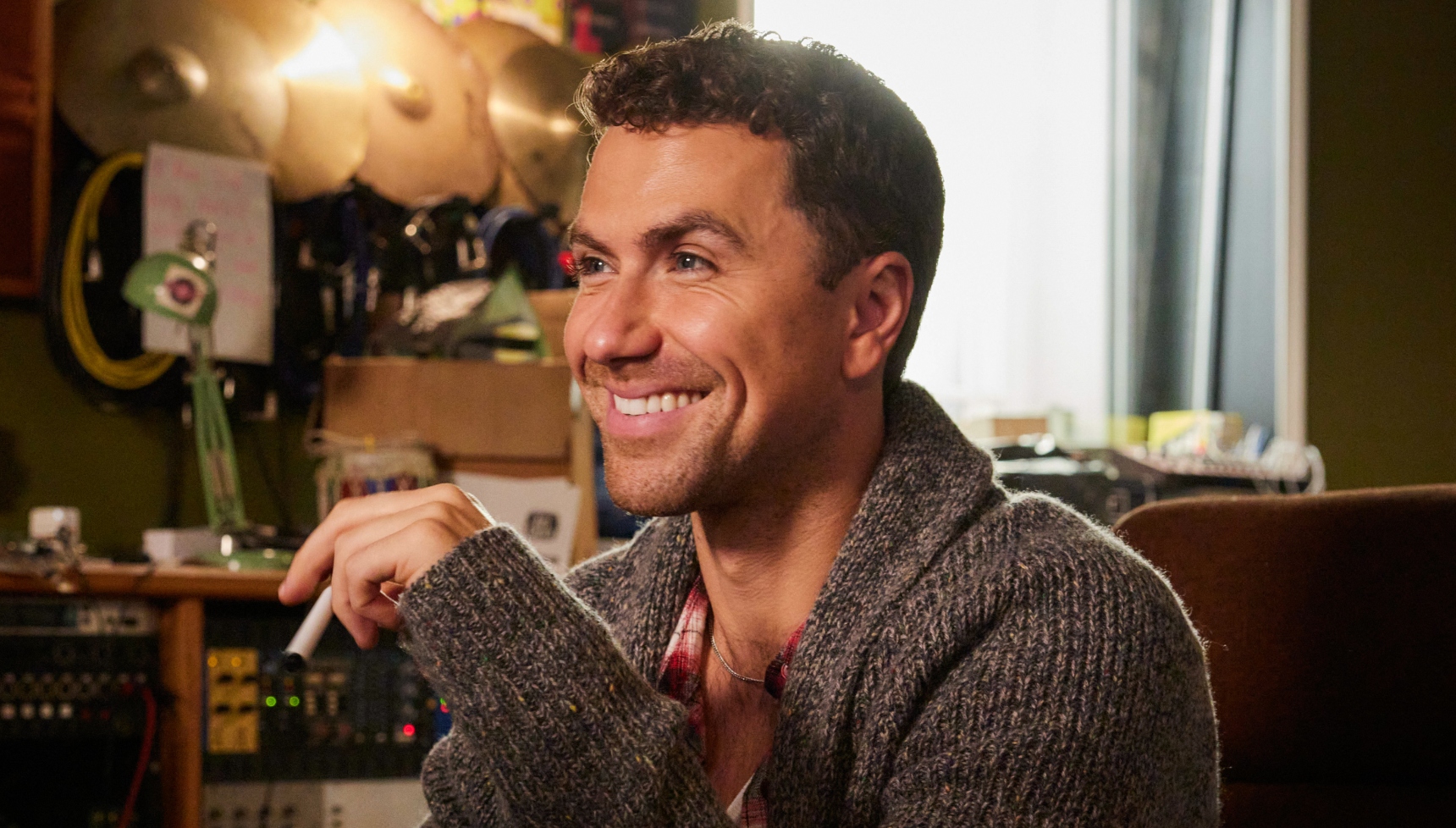 Richard Fleeshman as Jojo wearing a brown jumper and smiling to someone off camera