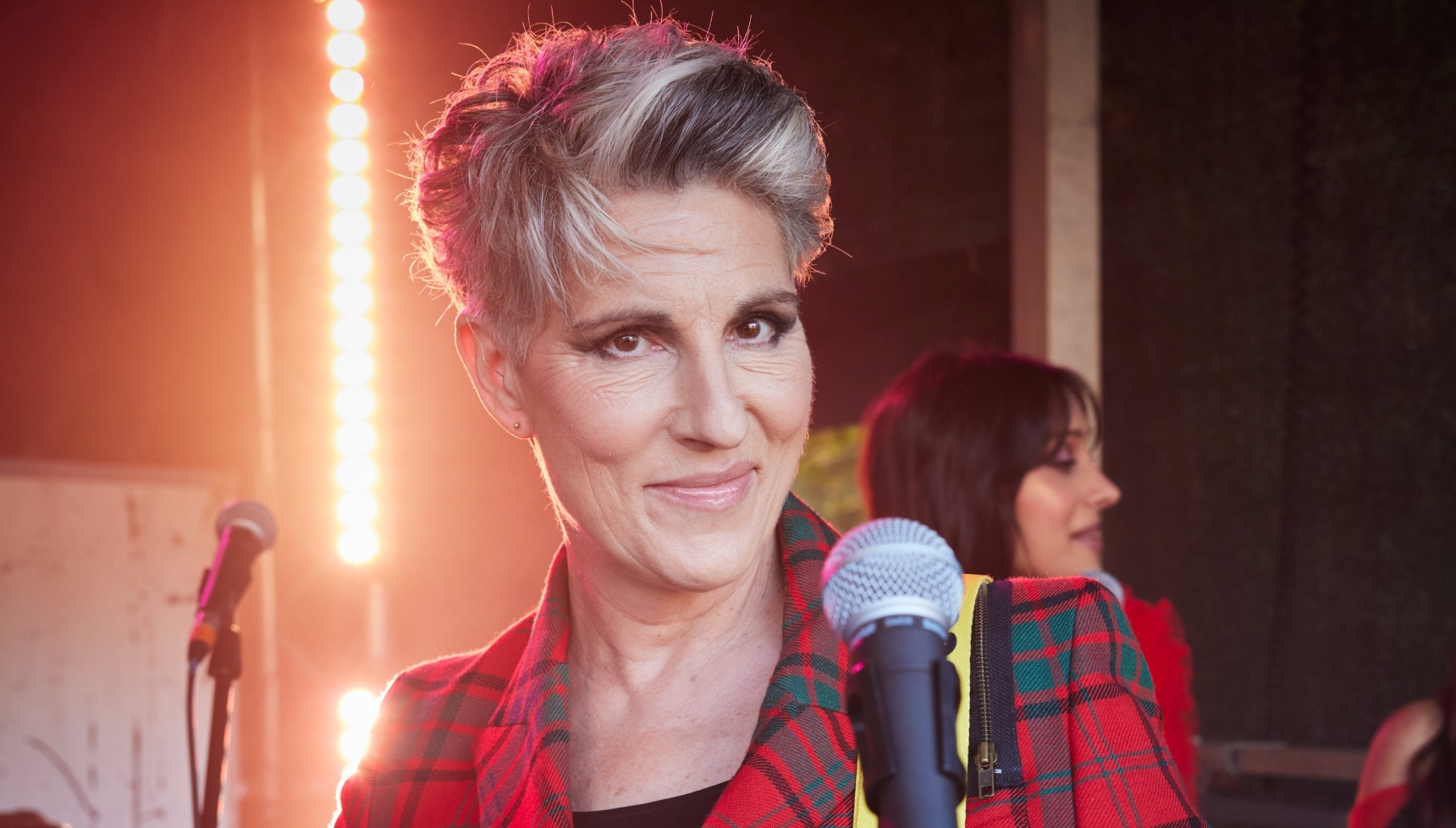 Tamsin Greig as Holly Gaskell wearing a tartan suit and standing in front of a microphone 