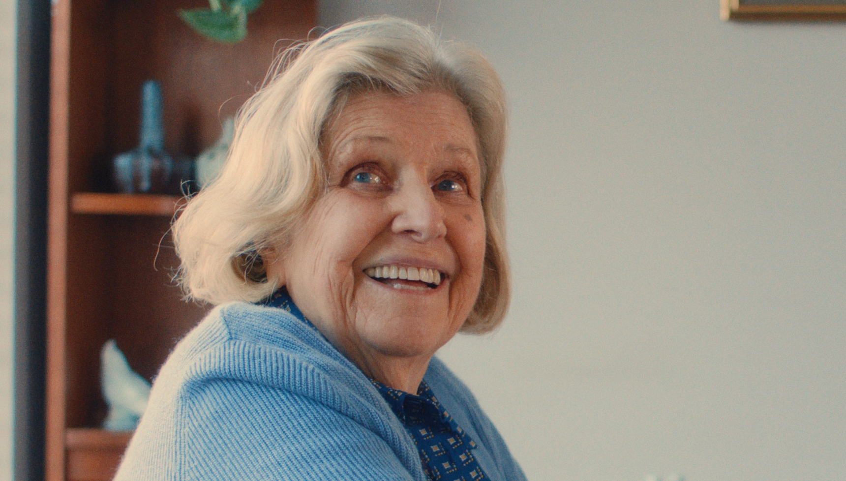 Anne Reid as Nancy wearing a blue cardigan and smiling 