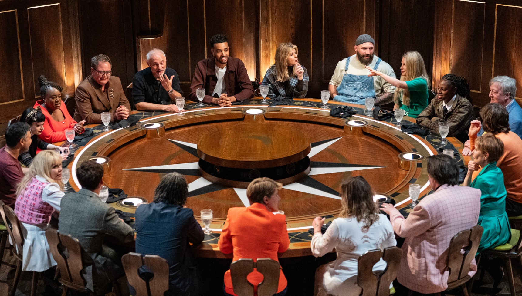 Multiple celebrities sat around a wooden table surrounded by atmospheric panelled walls