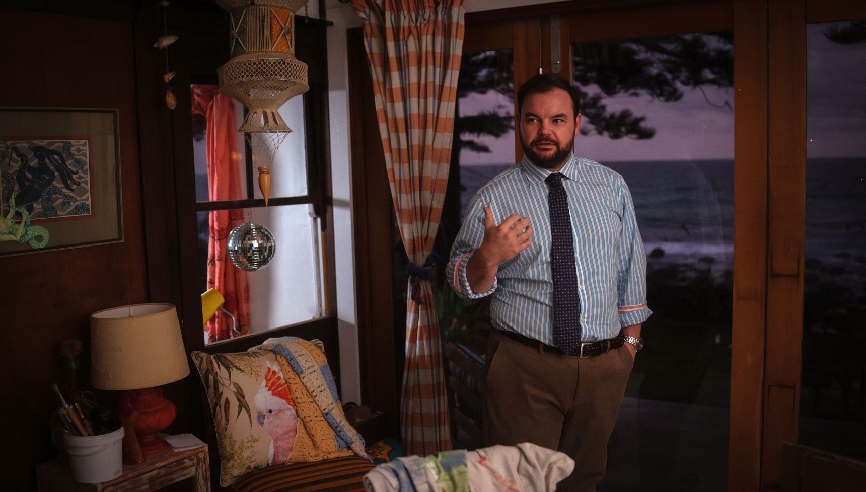 Colin standing in Mackenzie's house with the sun setting over the sea behind him