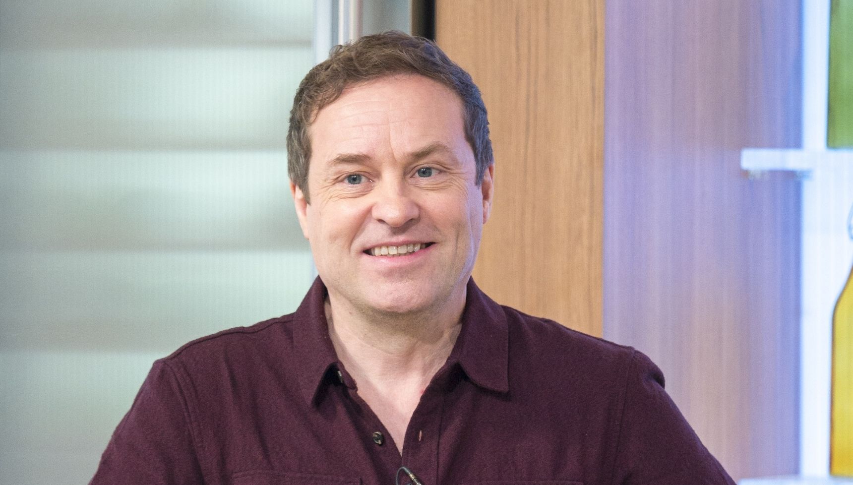 Ardal O'Hanlon in a dark red shirt smiling on a TV set