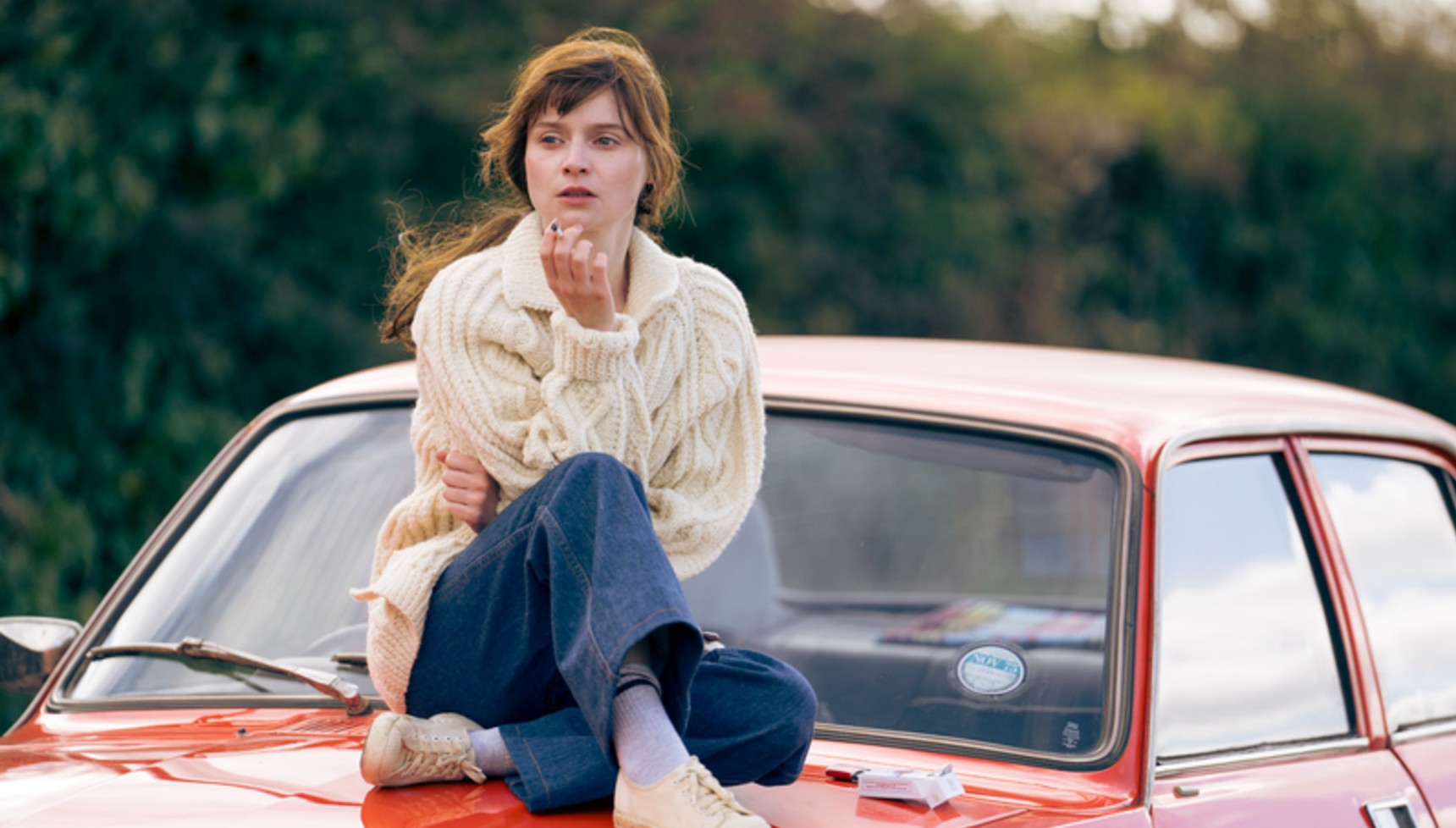 Woman smokes a cigarette while perched on the bonnet of a red car
