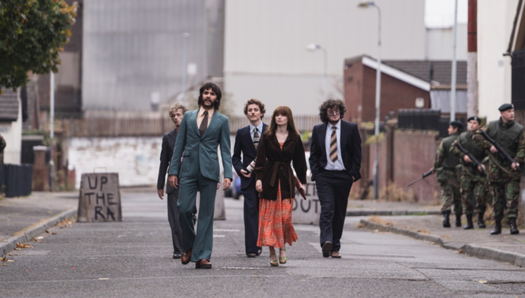 Five friends flanked by soldiers carrying guns walk down the street