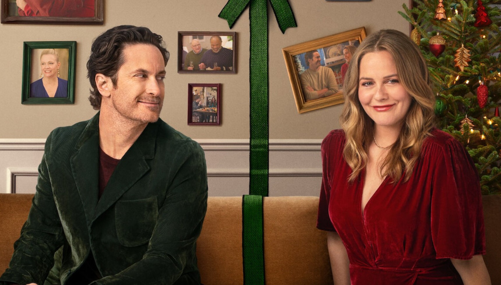 Oliver Hudson sitting on a couch with Alicia Silverstone next to a Christmas tree with a green bow between them