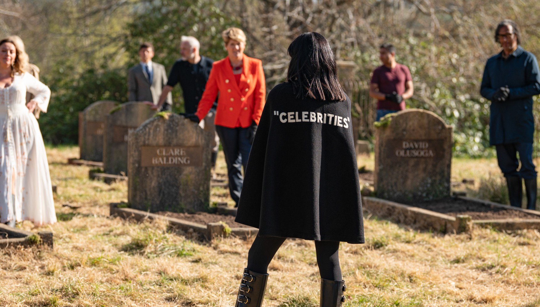 A group of celebrities stand around a graveyard in the daytime