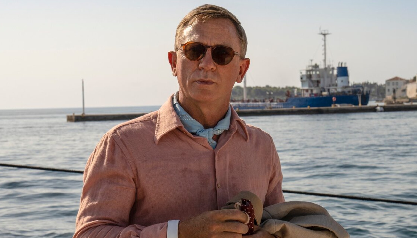 Daniel Craig wearing a pink shirt and sunglasses as he stands near the sea in Glass Onion