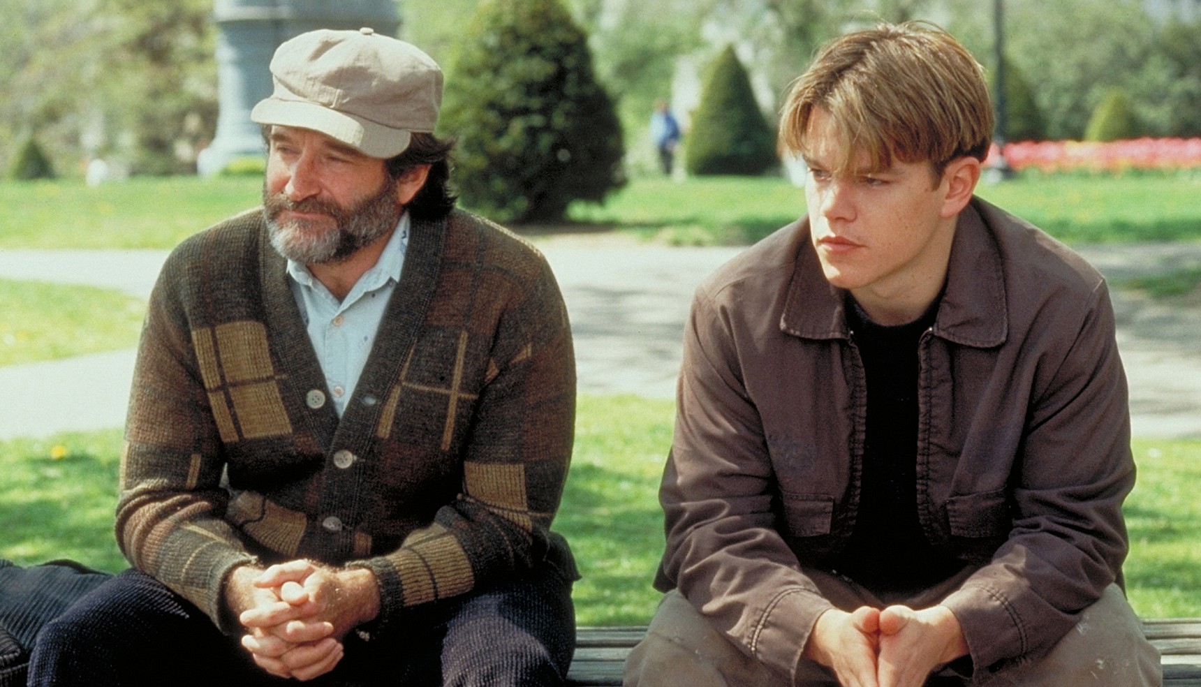 Robin Williams in a hat and Matt Damon sitting on a park bench together in Good Will Hunting