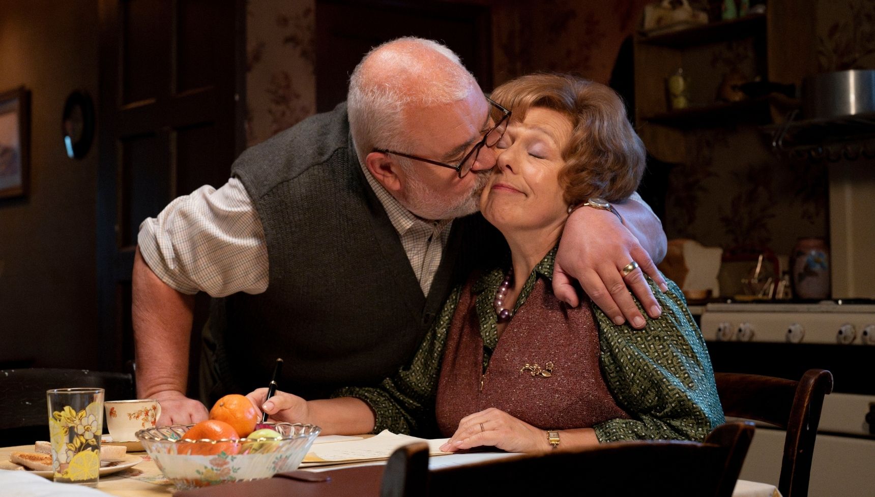 Fred kissing Violet on the cheek in their kitchen