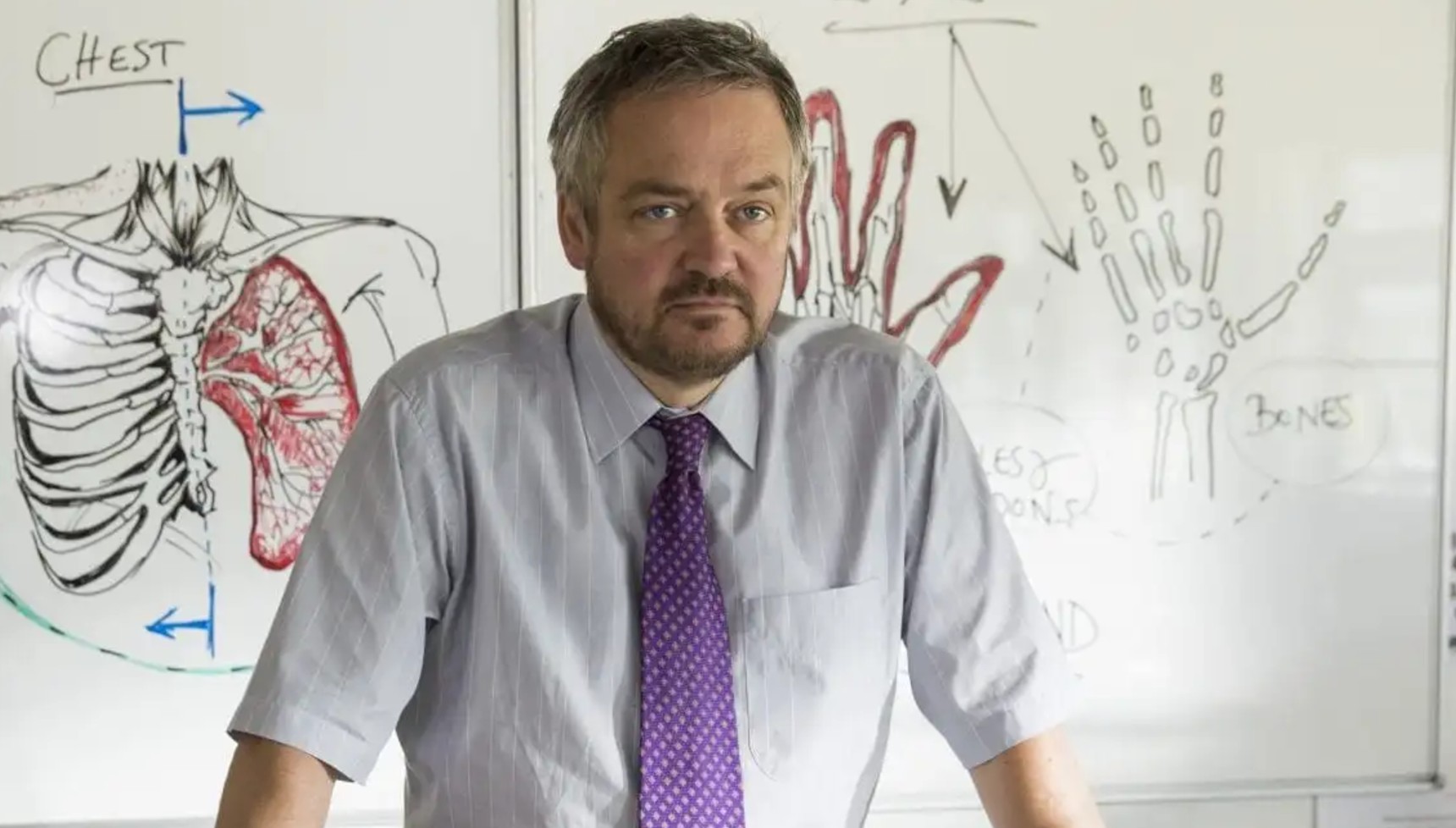 Charlie Higson wearing a purple tie in front of a whiteboard in Broadchurch