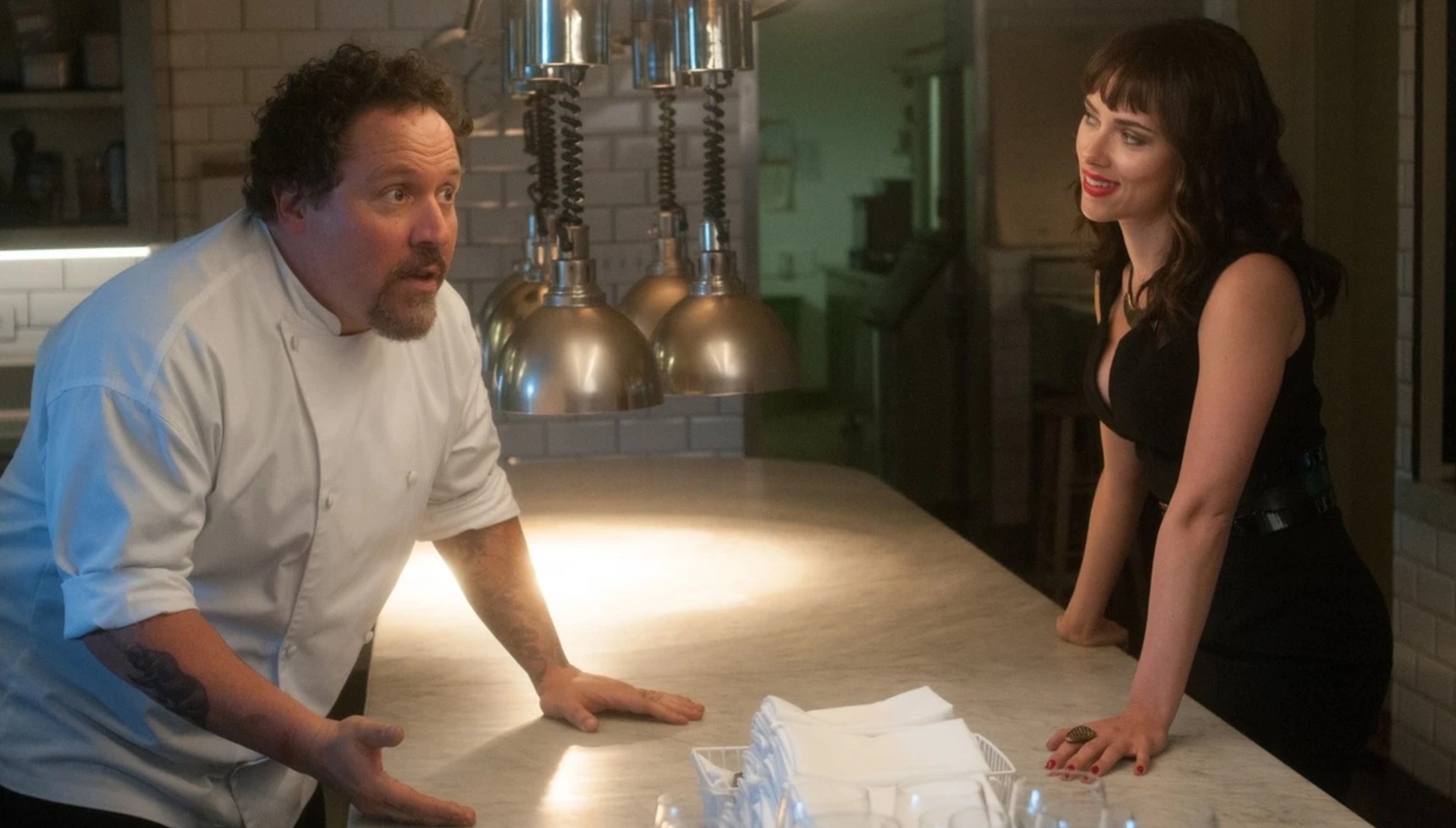 Jon Favreau in a chef's suit and Scarlett Johansson leaning on a table in a restaurant 