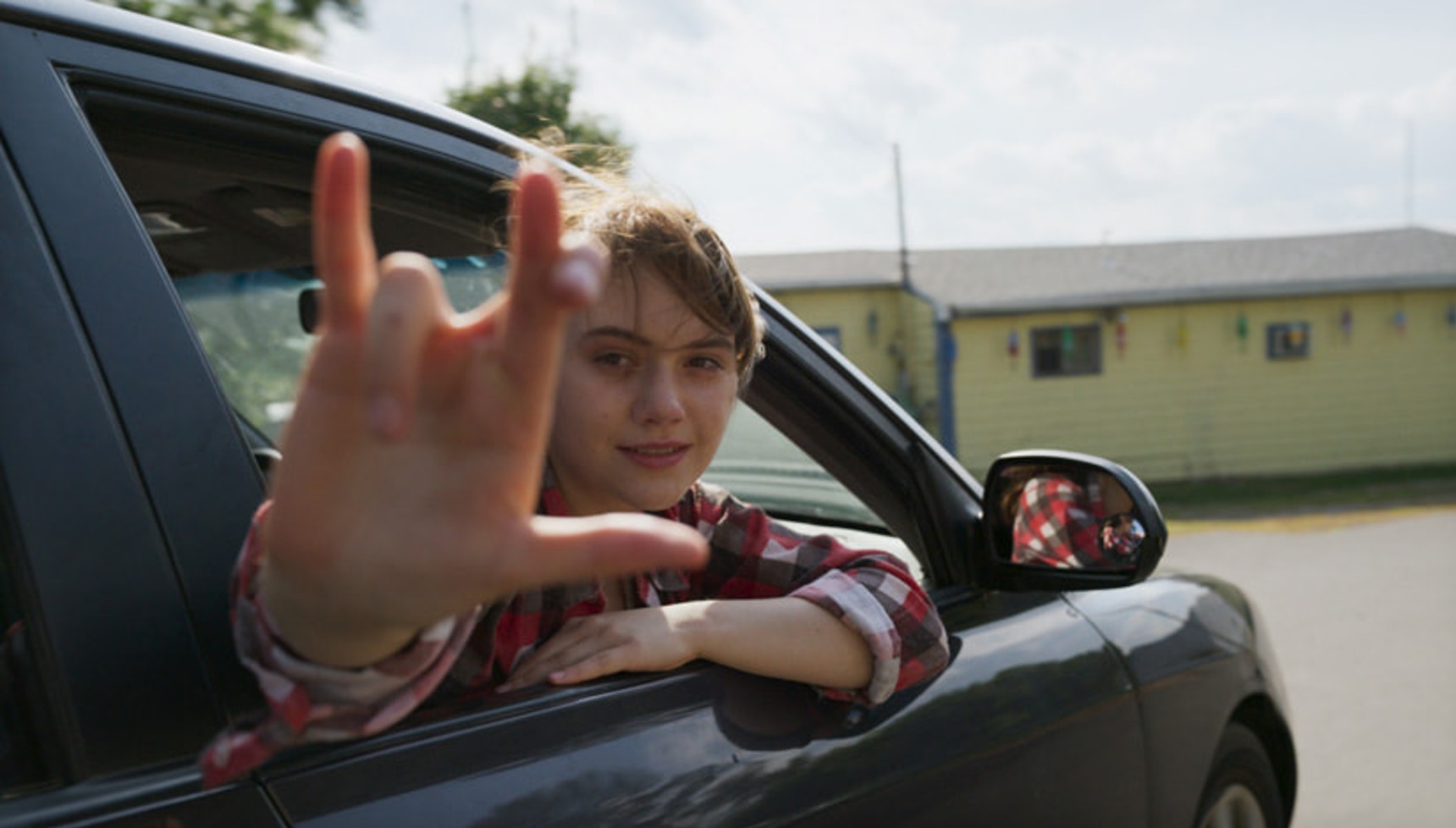 A girl hangs out of a car window gesturing with her fingers