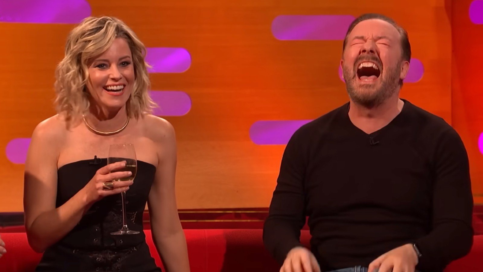 Elizabeth Banks holding a glass of red wine as Ricky Gervais laughs next to her on The Graham Norton Show