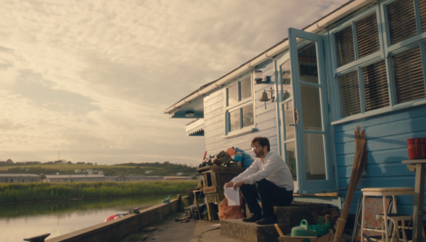 David Tennant sitting on the step outside a riverside blue chalet in Broadchurch