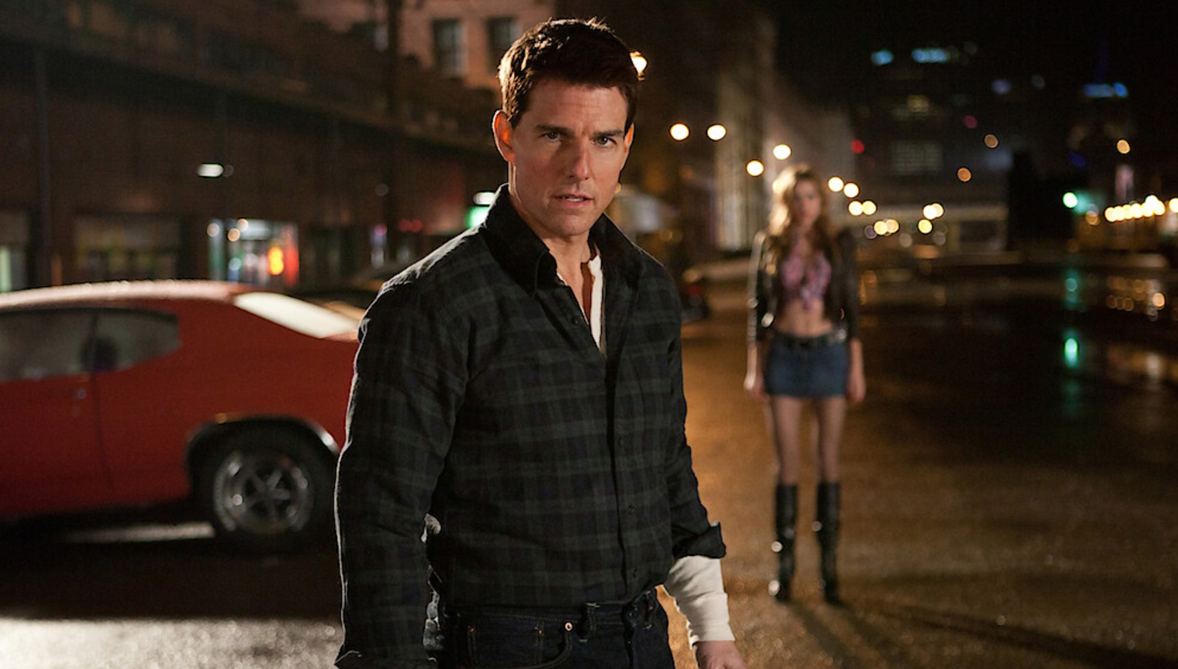 Tom Cruise with a bandage on his arm standing in front of a muscle car and a young woman