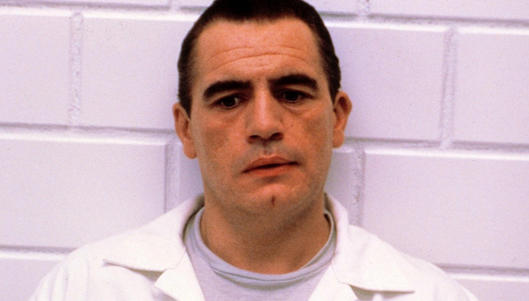 Brian Cox in a white shirt sitting against a white brick wall in Manhunter