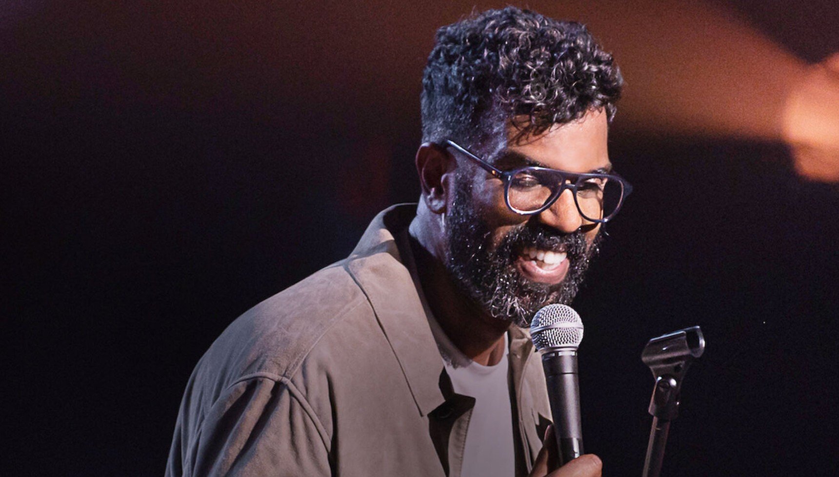 Romesh Ranganathan holding a microphone as he laughs