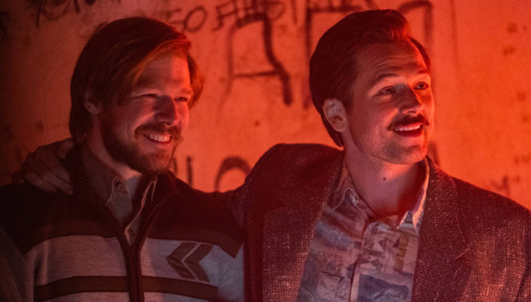 One man puts his arm around another as they smile