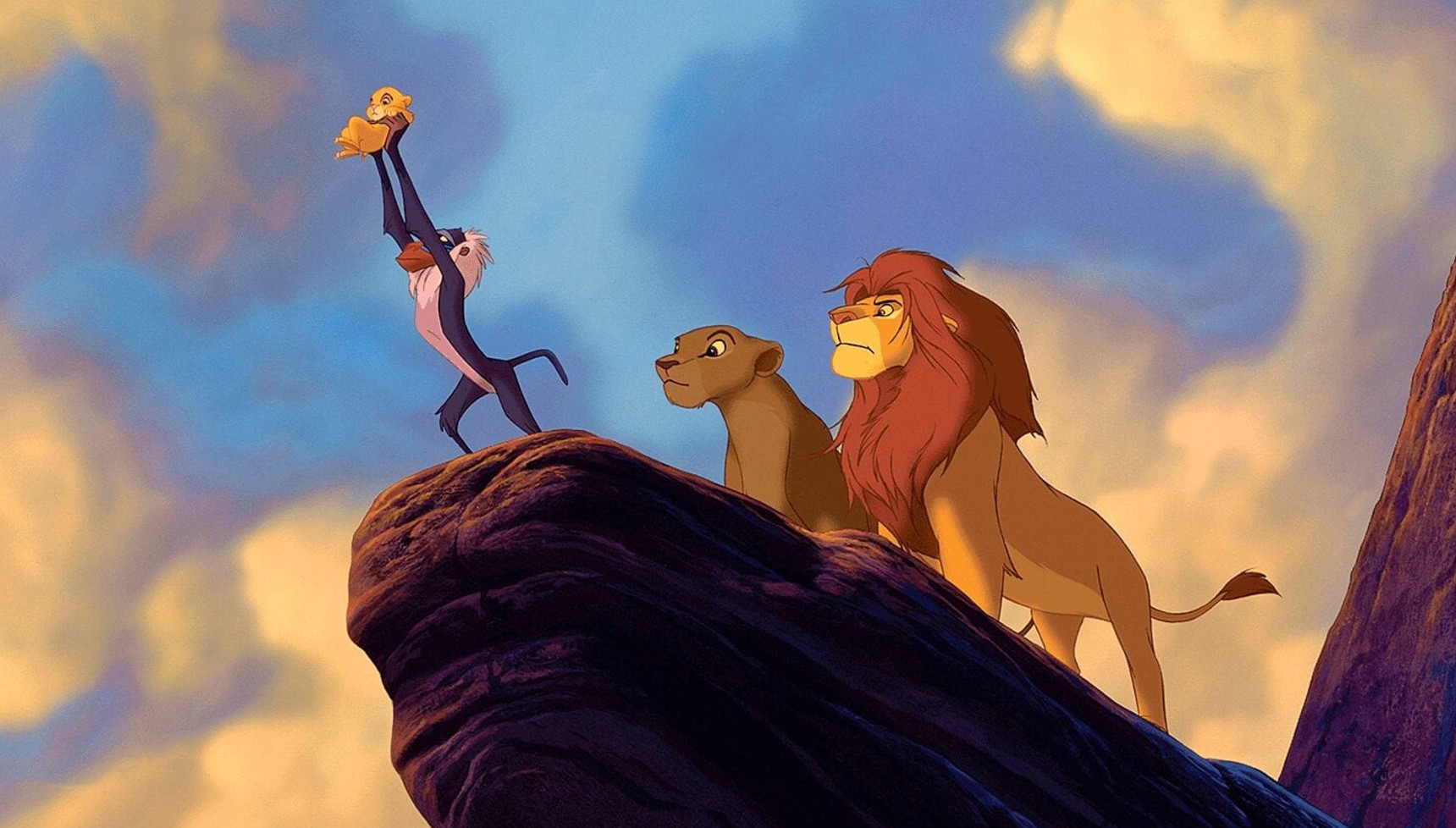 A monkey holding a lion cub in the air on a cliff as two lions stand behind him in The Lion King
