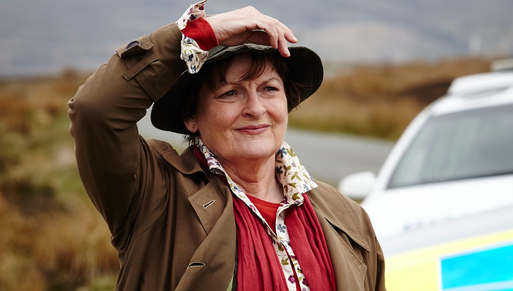 Brenda Blethyn in costume as Vera in front of a police car in Vera