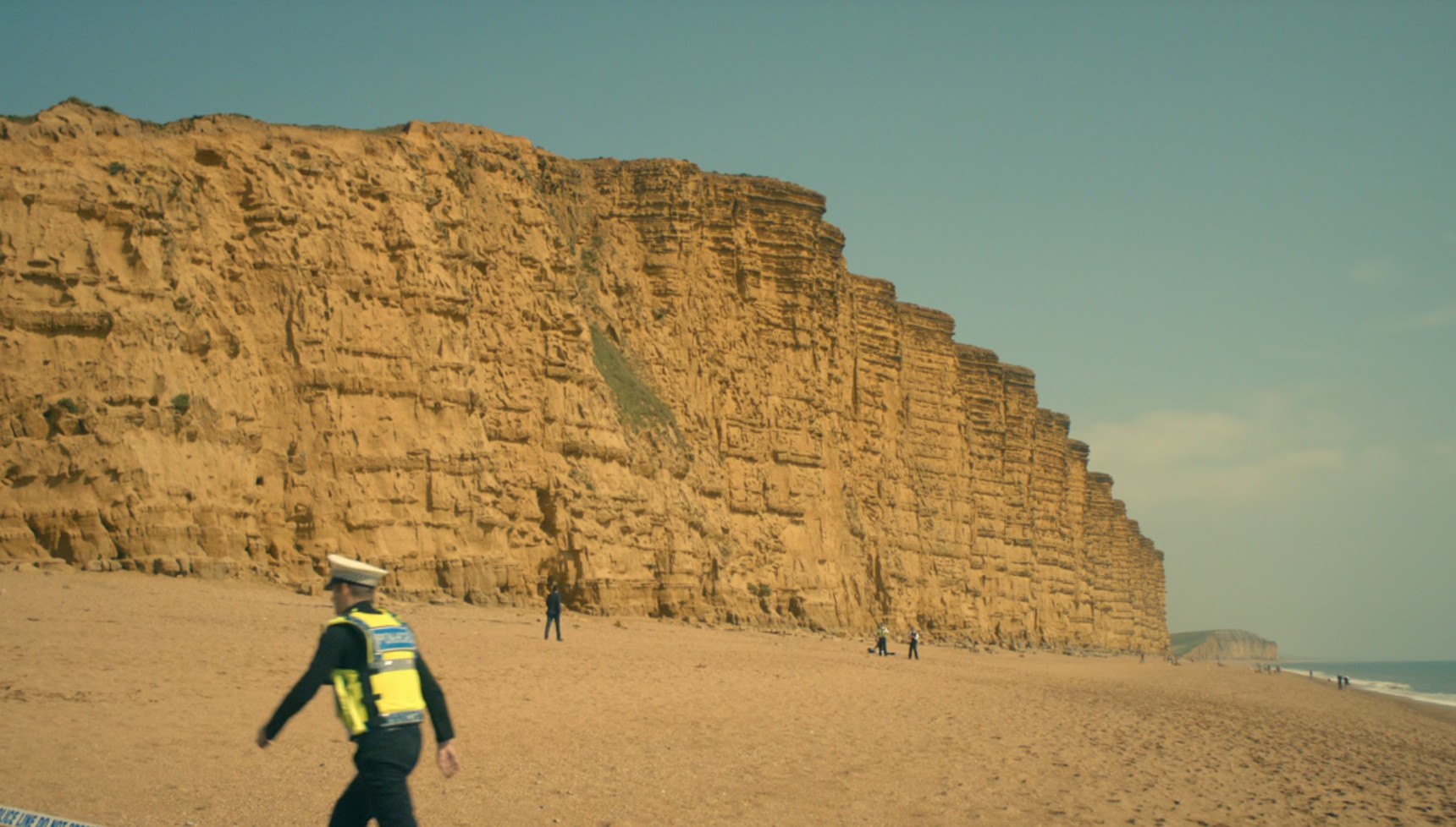 A police officer walking and Alec Hardy in the distance on the beach from Broadchurch