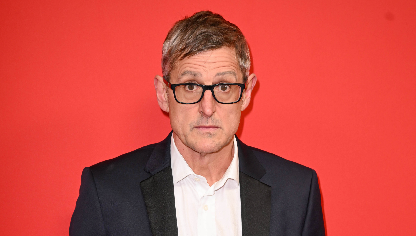 Louis Theroux standing against a red background