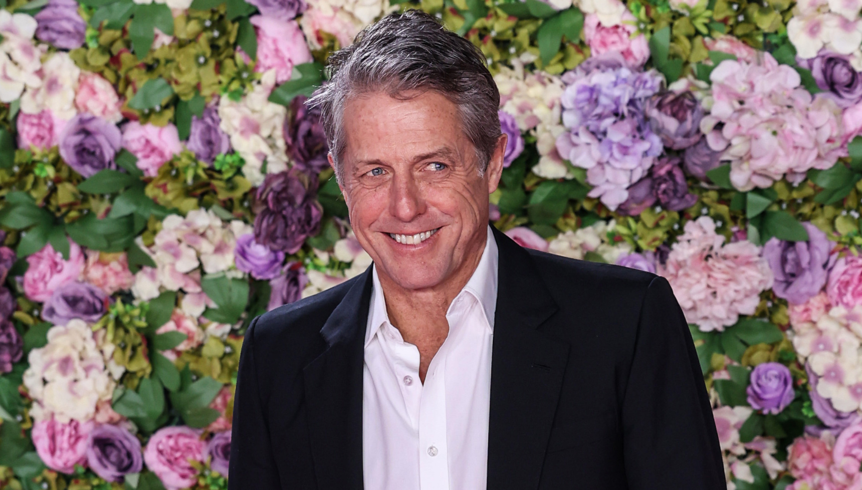 Hugh Grant smiling against a flower wall