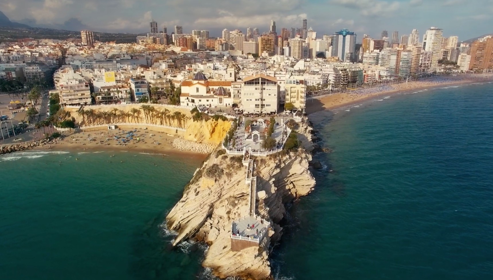 Panoramic shots show the rocky headland of the Balcón del Mediterráneo, a famous viewpoint in the city's Old Town. The sandy area to the left is the Cala del Mal Pas beach, nestled between the viewpoint and the port.